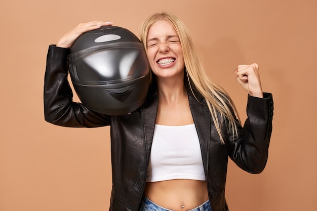 Retrato de jinete femenino joven alegre lleno de alegría con tirantes de dientes y pelo largo y recto posando aislado en chaqueta de cuero negro apretando el puño después de ganar la carrera