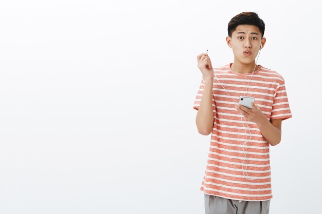 Retrato de un joven adolescente asiático impresionado y guapo con auriculares nuevos que se quitan los auriculares para expresar el asombro y la alegría de escuchar música a través del teléfono inteligente
