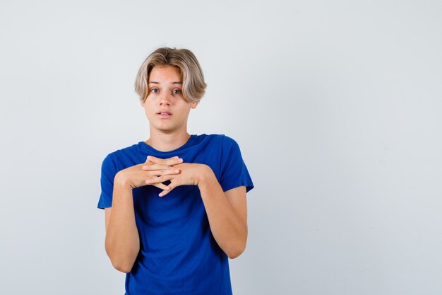 Retrato de joven adolescente manteniendo los dedos entrelazados sobre el pecho en camiseta azul y mirando desorientado vista frontal