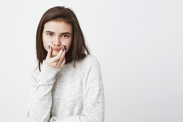 Retrato de joven adolescente sombrío expresando insatisfacción sosteniendo la boca con las manos para imitar la sonrisa