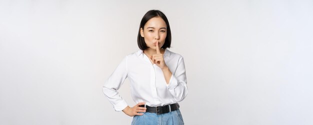 Retrato de una joven adulta asiática que se calla y dice calla shh presiona el dedo en los labios compartiendo el secreto no hables gesto tabú de pie sobre fondo blanco