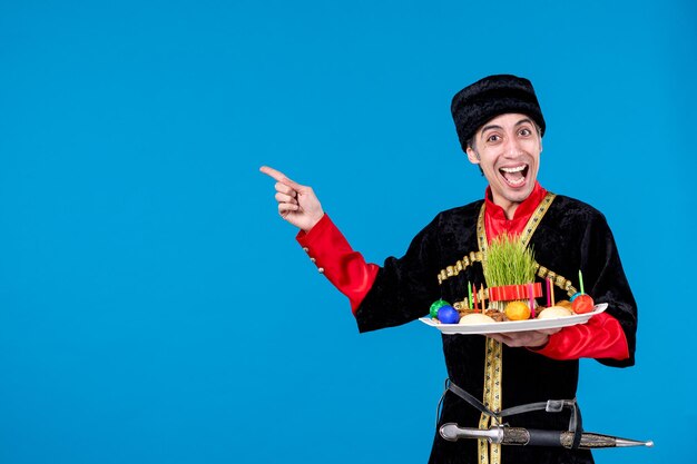 Retrato de un joven adulto sonriente y feliz con traje tradicional que muestra una bandeja llena de dulces nacionales que señalan algo en el fondo de la ola azul