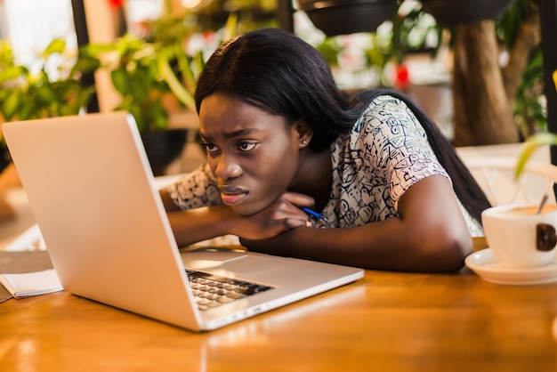 Retrato de una joven africana cansada sentada a la mesa con el ordenador portátil mientras duerme en un café