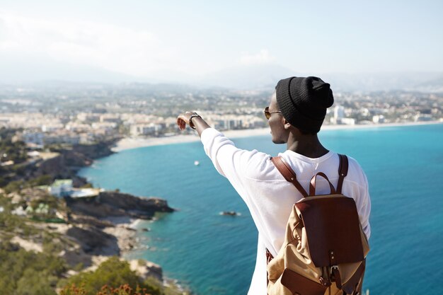 Retrato de un joven africano irreconocible con sombrero negro inconformista de pie en la plataforma de turismo admirando el mar y la hermosa ciudad turística apuntando con el dedo a lugares lejanos que visitará