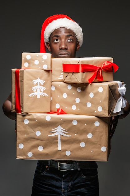 El retrato del joven africano negro guapo sorprendido con sombrero de Santa con regalos sobre fondo oscuro. Emociones humanas positivas y concepto de feliz Navidad