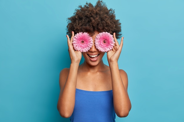 El retrato de una joven afroamericana feliz cubre los ojos con gerberas rosadas, se divierte, sostiene sus flores favoritas, tiene una sonrisa con dientes, se viste con un top azul, disfruta del tiempo libre.