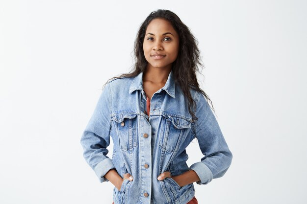 Retrato de joven afroamericana con piel oscura con chaqueta vaquera y camiseta roja con expresión seria, de pie con las manos en los bolsillos. Modelo femenino de piel oscura con d