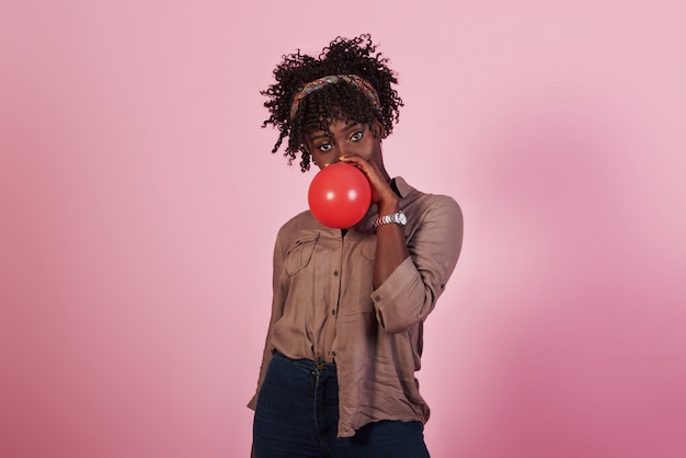 Retrato de joven afroamericana soplando globo rojo ar fondo rosa studio
