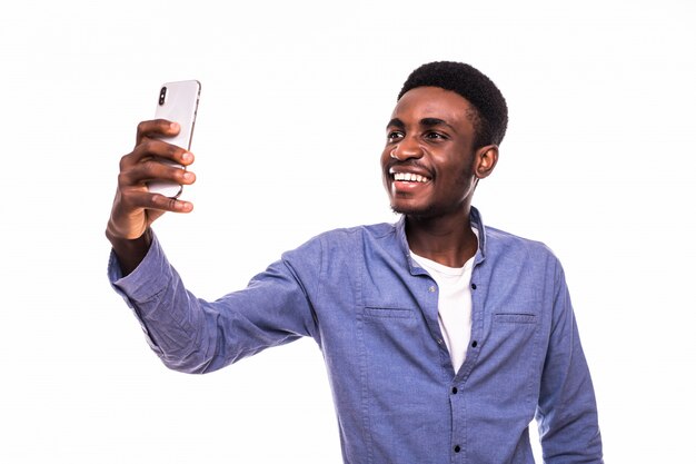 Retrato de joven afroamericano guapo con smartphone para tomar fotos selfie y sonriente de pie contra la pared blanca