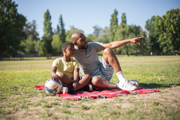 Retrato de joven afroamericano y niño descansando en el suelo. Papá feliz y su hijo sentados en una manta, papá mostrando objeto con su dedo, ambos mirando allí. Concepto de ocio y actividad de verano