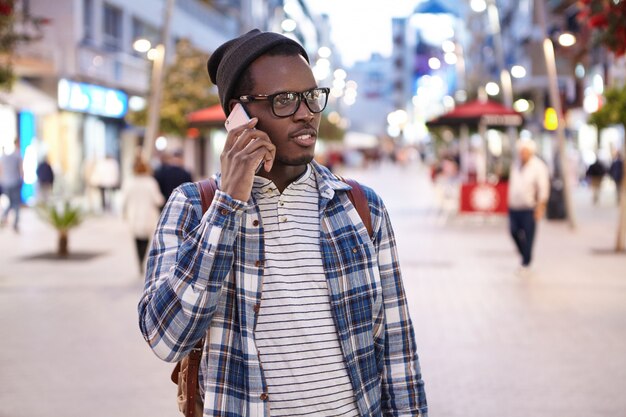 Retrato de joven afroamericano vistiendo ropa elegante y accesorios hablando por teléfono inteligente en su camino a casa