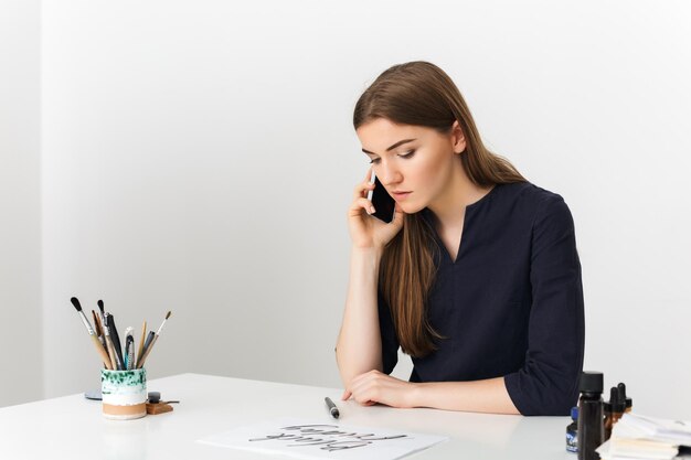 Retrato de una joven agradable sentada en el escritorio blanco y hablando por su teléfono celular mientras miraba soñadoramente el papel aislado