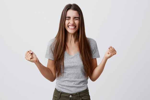 Retrato de joven alegre feliz hermosa estudiante femenina con cabello largo y oscuro con atuendo informal gris sonriendo con dientes, extendiendo las manos con los ojos vestidos, siendo extremadamente feliz finalmente obtener un papel en la película