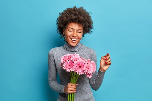 Retrato de joven alegre mujer afroamericana con cabello rizado tiene ramo de gerberas rosas sonríe ampliamente