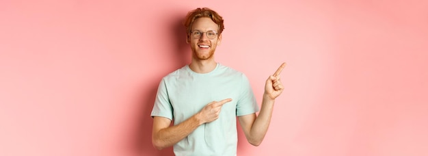 Foto gratuita retrato de un joven alegre con el pelo rojo y gafas señalando con el dedo en la esquina superior izquierda a