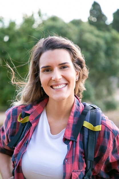 Retrato de joven alegre posando, sonriendo. Hermosa mujer caucásica con mochila, de pie y disfrutando de la naturaleza. Concepto de turismo, aventura y vacaciones de verano.