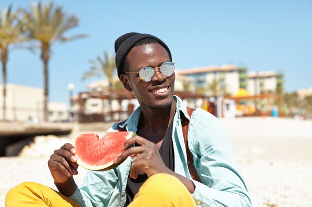 Retrato de joven alegre relajante en la playa urbana sosteniendo una rodaja de sandía madura