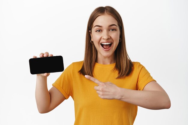 Retrato de una joven alegre señalando con el dedo a la venta, pantalla de teléfono inteligente horizontal, interfaz o aplicación en exhibición, de pie contra el fondo blanco