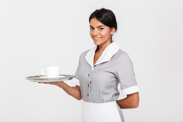 Retrato de joven alegre en uniforme gris con bandeja de metal con una taza de café