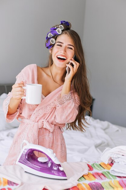 Retrato joven ama de casa con pelo largo en bata de baño rosa y rizador en la cabeza a planchar en casa. Ella sostiene una taza, hablando por teléfono y riendo.