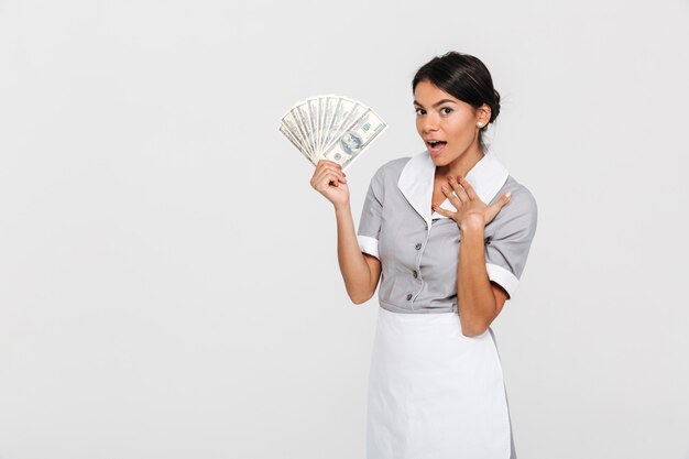 Retrato de joven ama de llaves asombrada en uniforme con abanico de billetes de dólar