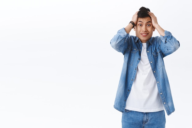 Foto gratuita retrato de joven ansioso avergonzado asiático agarra la cabeza en pánico, sacudiéndola en negación, se siente frustrado y nervioso, tiene un gran problema, de pie pared blanca preocupada