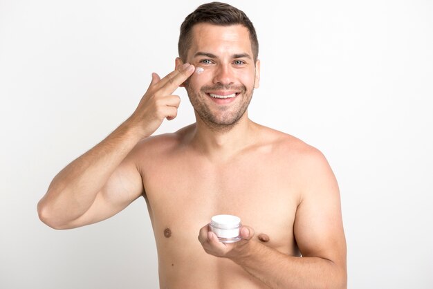 Retrato de joven aplicando crema para la cara en la cara de pie contra el fondo blanco.