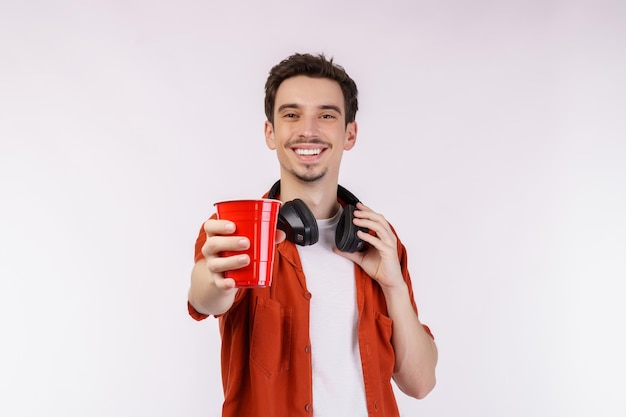 Retrato de un joven apuesto con auriculares de pie y mostrando café mientras mira a la cámara con fondo blanco aislado