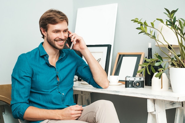 Foto gratuita retrato de joven apuesto hombre de negocios. hombre pensativo vestido con camisa de jeans azul. modelo barbudo posando en la oficina cerca del escritorio de papel y hablando por teléfono.