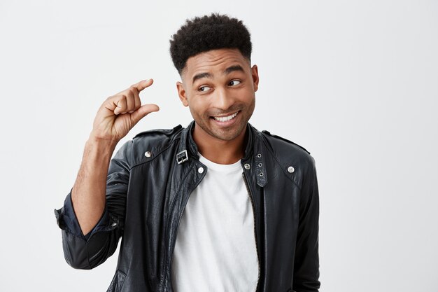 Retrato de joven apuesto hombre de piel oscura y divertido con peinado afro en camiseta blanca y chaqueta de cuero gesticulando con la mano, mostrando poco tamaño mirando a un lado con expresión cínica de la cara.