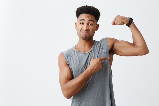 Retrato de joven apuesto hombre de piel oscura con peinado afro en camisa sin mangas gris que muestra los músculos, señalando con expresión de confianza en sí mismo.