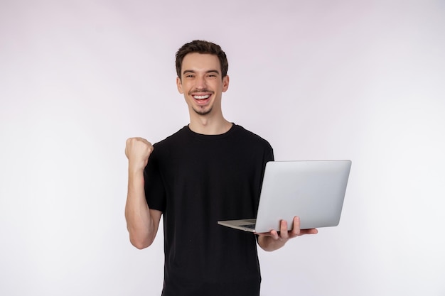 Foto gratuita retrato de un joven apuesto hombre sonriente sosteniendo una laptop en las manos escribiendo y navegando por páginas web mientras hace un gesto de puño cerrado ganador aislado en fondo blanco