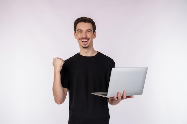 Retrato de un joven apuesto hombre sonriente sosteniendo una laptop en las manos escribiendo y navegando por páginas web mientras hace un gesto de puño cerrado ganador aislado en fondo blanco