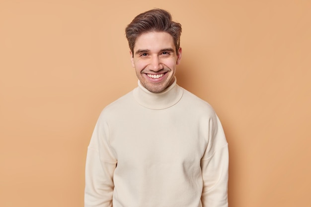 Retrato de un joven apuesto con una sonrisa dentuda de pelo oscuro que mira alegremente a la cámara y viste un cuello de tortuga blanco casual aislado sobre un fondo marrón Sonriente modelo masculino adulto de buen humor