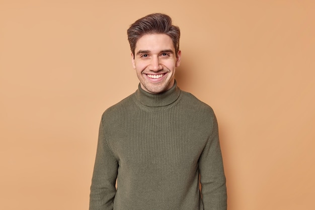 Retrato de un joven apuesto con sonrisas de cabello oscuro expresa felizmente emociones positivas vestido con un cuello de tortuga casual escucha buenas nes aislado sobre un fondo marrón contento de conocer al viejo mejor amigo