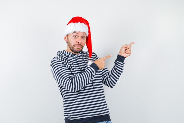 Retrato de joven apuntando a la esquina superior derecha con capucha, gorro de Papá Noel y mirando desconcertado