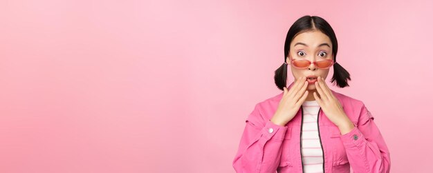 Retrato de una joven asiática con elegantes gafas de sol que parece una reacción de incredulidad sorprendida de pie sobre un fondo rosa