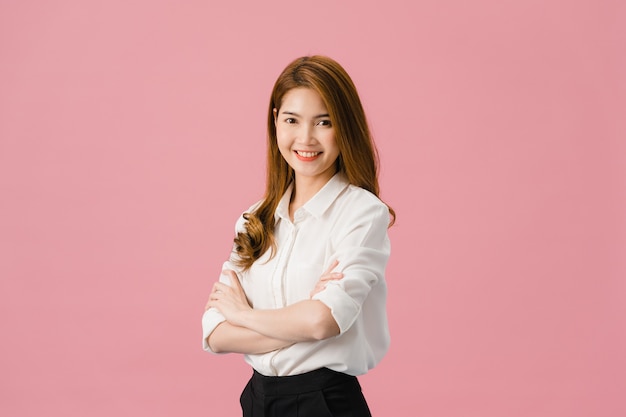 Retrato de joven asiática con expresión positiva, brazos cruzados, sonrisa amplia, vestida con ropa casual y mirando a cámara sobre fondo rosa.