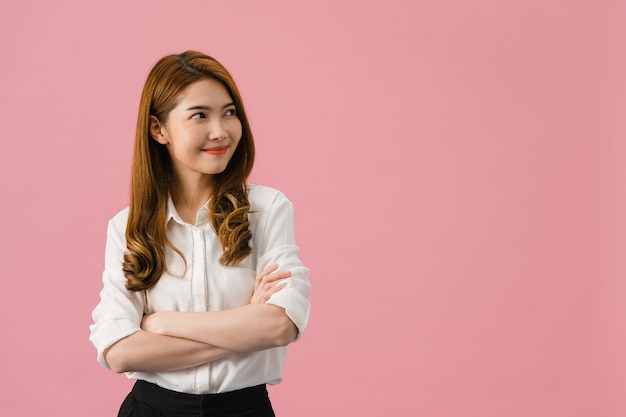 Retrato de joven asiática con expresión positiva, brazos cruzados, sonrisa amplia, vestida con ropa casual y mirando el espacio sobre fondo rosa.