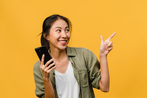 Retrato de joven asiática que usa un teléfono móvil con expresión alegre, muestra algo sorprendente en el espacio en blanco en ropa casual y se para aislado sobre una pared amarilla. Concepto de expresión facial.