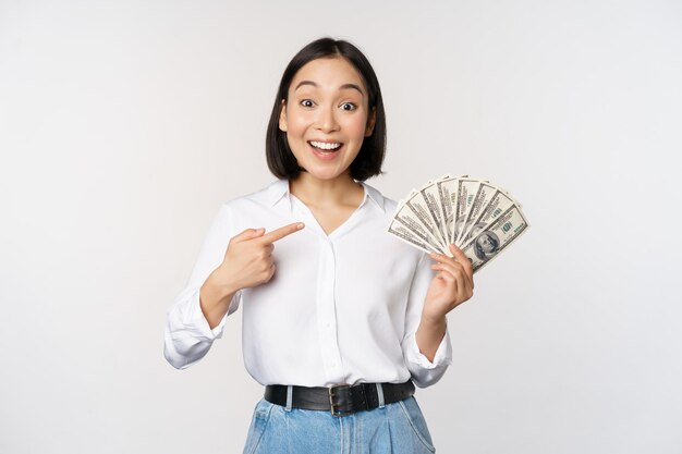 Retrato de una joven asiática señalando sus dólares de dinero mostrando dinero en efectivo de pie sobre fondo blanco.