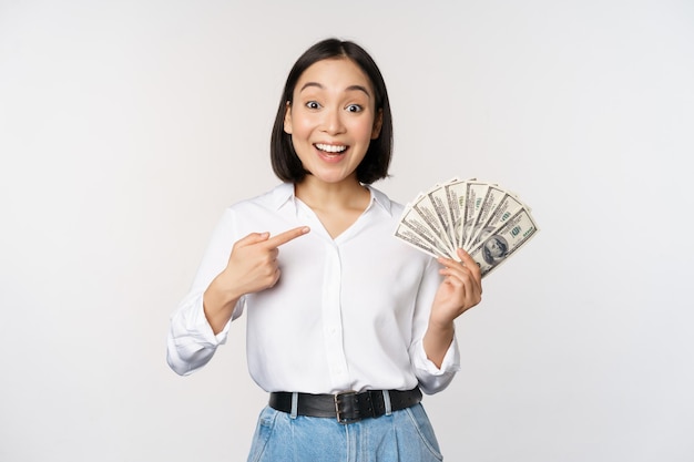 Retrato de una joven asiática señalando sus dólares de dinero mostrando dinero en efectivo de pie sobre fondo blanco.