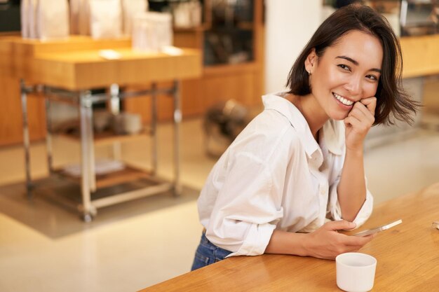 Retrato de una joven asiática sentada en un café sosteniendo un teléfono inteligente chateando y enviando mensajes mientras bebe