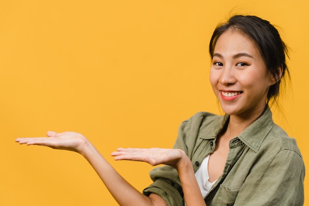 Foto gratuita retrato de joven asiática sonriendo con expresión alegre, muestra algo sorprendente en el espacio en blanco en un paño casual aislado sobre la pared amarilla. concepto de expresión facial.