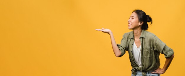 Retrato de joven asiática sonriendo con expresión alegre, muestra algo sorprendente en el espacio en blanco en ropa casual y de pie aislado sobre una pared amarilla. Banner panorámico con espacio de copia.