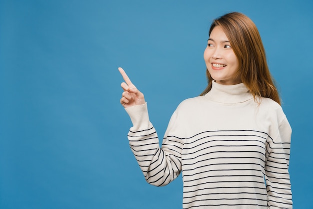 Retrato de joven asiática sonriendo con expresión alegre, muestra algo sorprendente en el espacio en blanco en ropa casual y de pie aislado sobre la pared azul