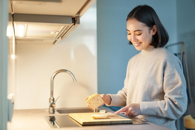 Retrato de una joven asiática sonriente parada en la cocina y haciendo un sándwich cocinando para ella