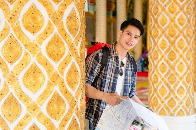 Retrato joven asiático mochilero macho de pie y comprobando la dirección en el mapa de papel en la mano en el hermoso templo tailandés y sonrisa