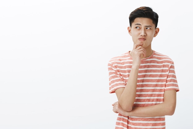 Retrato de un joven asiático con problemas tratando de pensar en un plan o una idea, de pie en una pose pensativa con la mano en la barbilla, mirando cuestionado y vacilante en la esquina superior izquierda