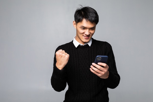 Retrato de un joven asiático se ve feliz mientras lee una buena noticia en el teléfono inteligente en la pared blanca